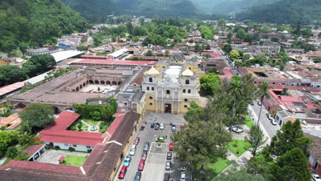 drone footage of iglesia de la merced church in antigua, guatemala on a bright cloudy day with colorful red town rooftops and green forest surrounding the city