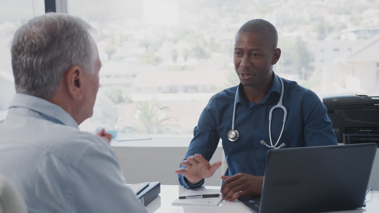 Senior Male Patient In Consultation Shaking Hands With Doctor Sitting At Desk In Office