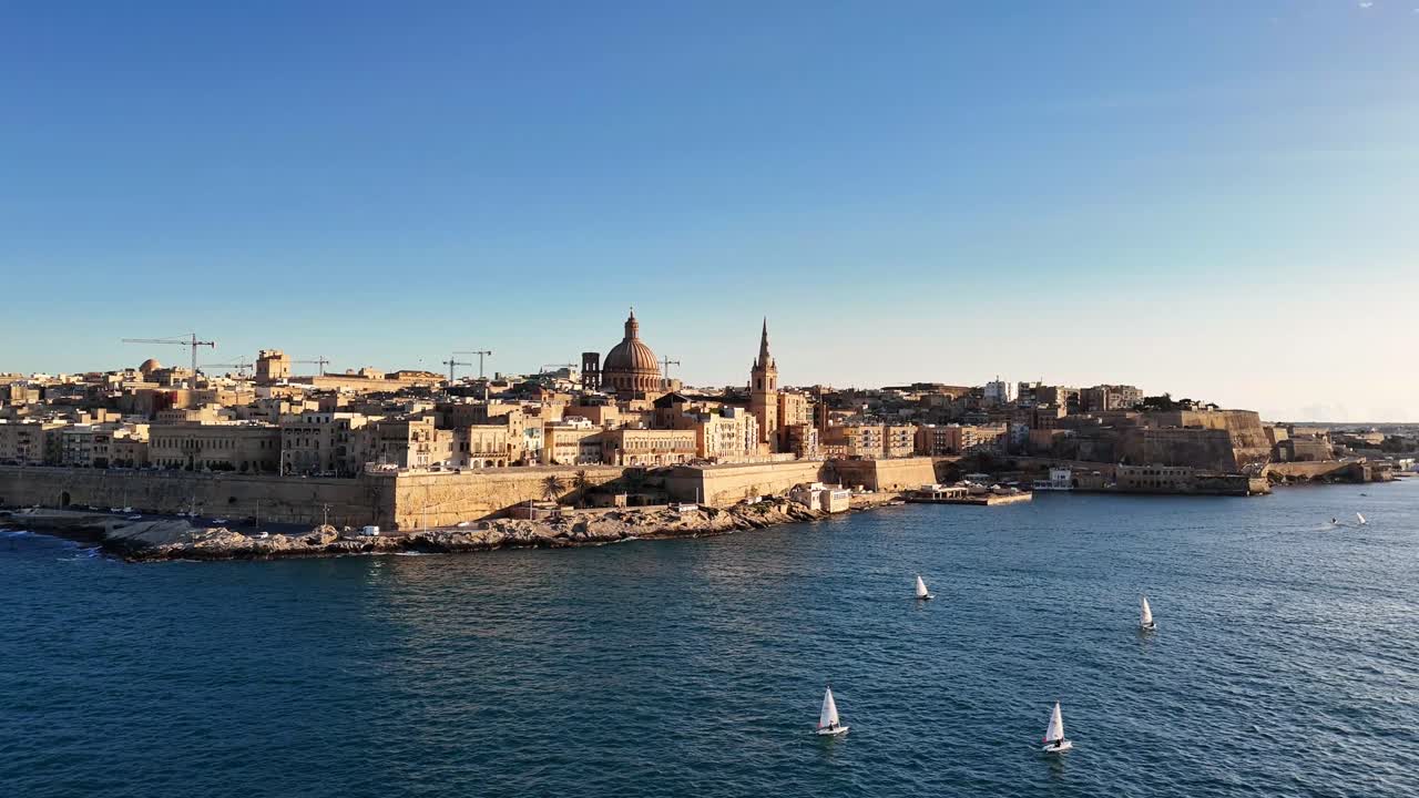 Ampia ripresa con drone della storica città di La Valletta a Malta, con la cupola della Basilica di Nostra Signora del Monte Carmelo e barche a vela nel porto mediterraneo al tramonto