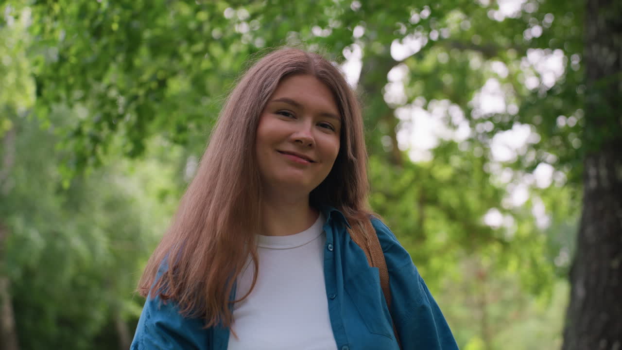 Una mujer hermosa sonríe con elegancia en un parque verde y vibrante, vestida con una camisa azul y una camiseta blanca, de pie bajo la luz del sol rodeada de árboles frondosos, mostrando una confianza tranquila.