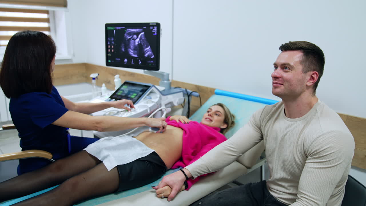 Happy Caucasian couple having pregnancy ultrasound examination. Doctor is focused on the monitor of ultrasonic machine.