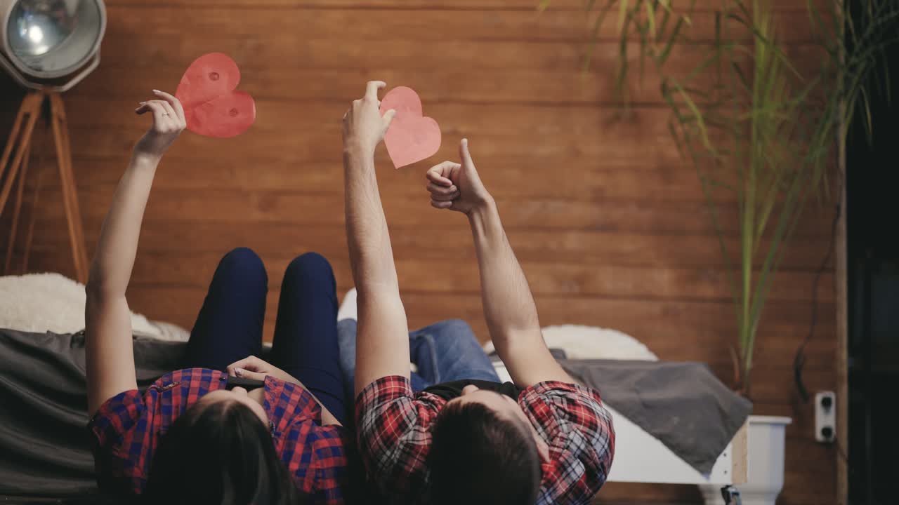 Romantic couple lying in bed and playing with red paper hearts. Man and woman hold their hands up with hearts in bedroom and show yes gesture. Valentine's day