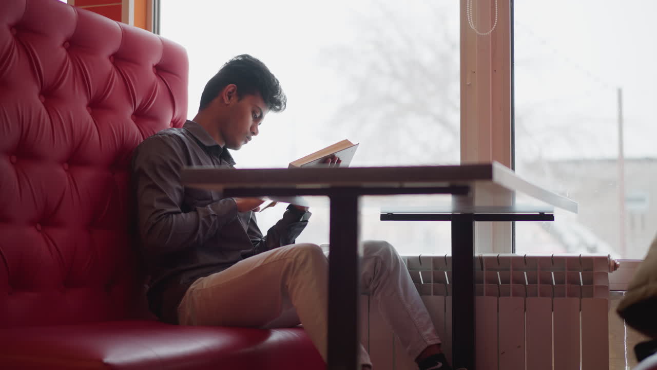 Young man sitting in cozy booth near window on snowy day reading book while using phone, relaxed mood, focused posture, natural light, casual clothes, quiet indoor winter atmosphere, peaceful scene