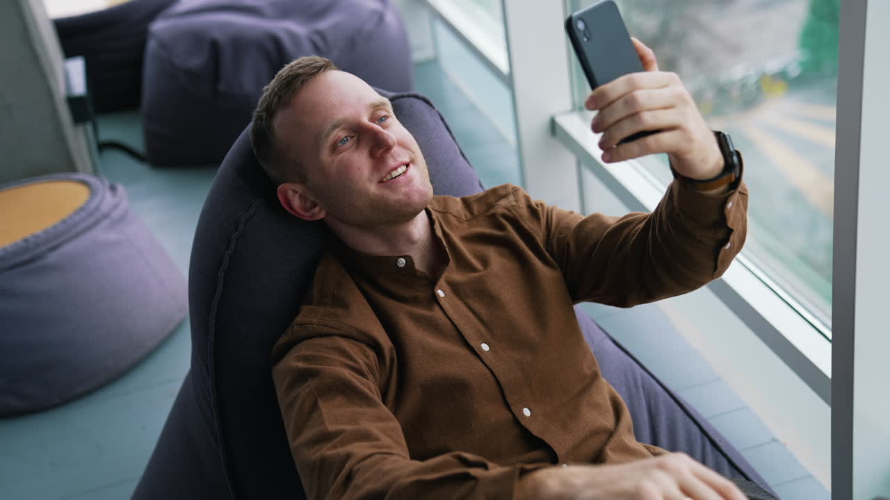 Man is making a video call. Handsome young businessman sitting in a soft chair near the window and talking through the phone in office.