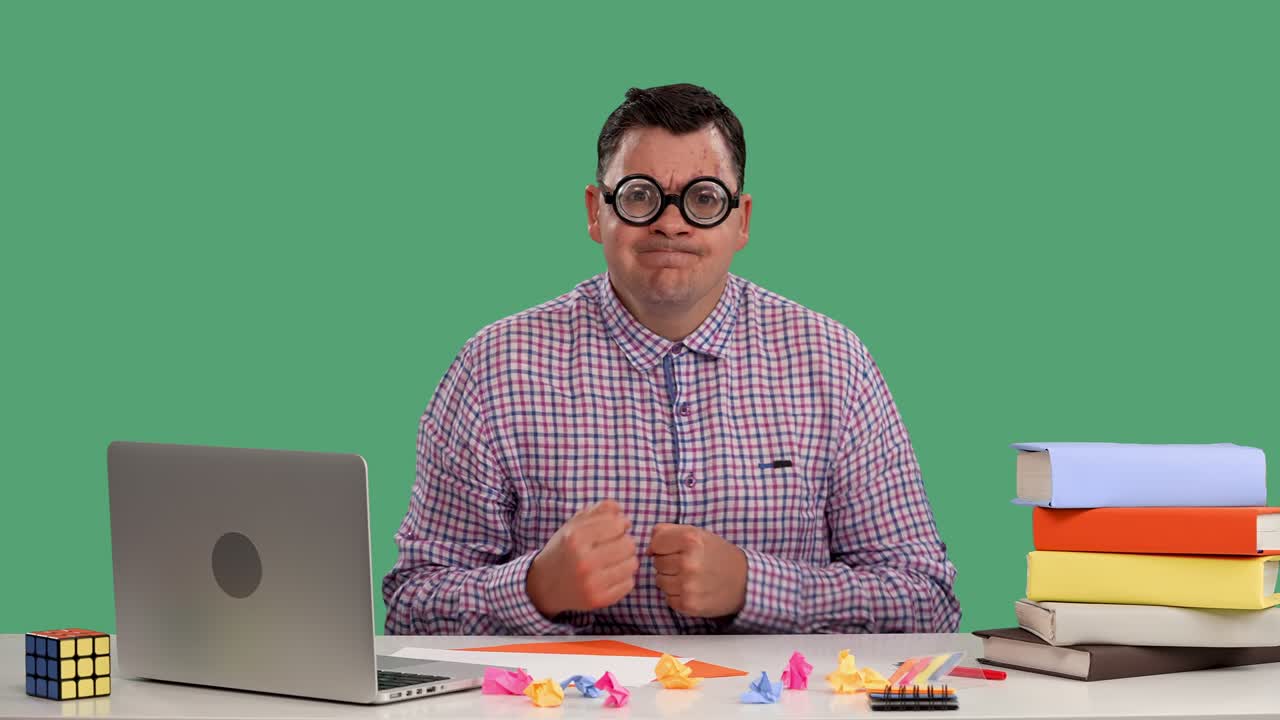 An angry man sits at a desk in front of a laptop and books, shows his fist to his interlocutor, hits the table with his fists. Portrait of a man with glasses on a green screen. Slow motion. Close up