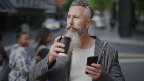 portrait of mature businessman using smartphone texting checking messages drinking coffee on city street waiting