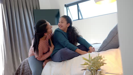 Happy-biracial-mother-talking-with-daughter-and-brushing-her-hair-in-sunny-bedroom