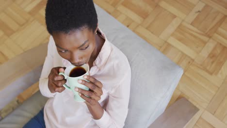 Happy-african-american-woman-sitting-on-armchair-in-living-room,-drinking-coffee