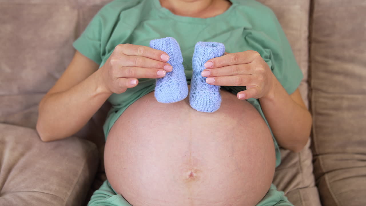 Beautiful pregnant woman with baby shoes at home. Pregnant woman preparing for motherhood, holding small baby shoes near belly