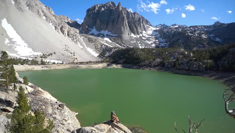 Young-Woman-in-Picturesque-Landscape,-Sitting-on-a-Rock-Above-Glacial-Lake-Under-Peak-With-Snow-on-Slopes