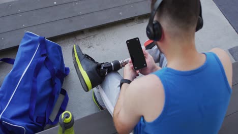 caucasian disabled male athlete with prosthetic leg sitting, wearing headphones, using smartphone