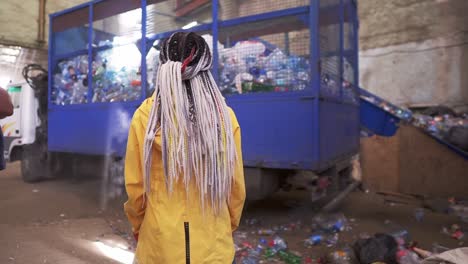 Backside-footage-of-a-woman-with-dreadlocks-wearing-yellow-jacket-watching-the-process-on-waste,-recycling-factory.-Big-truck-with-cage-carcase-of-used-plastic-bottles-for-recycling