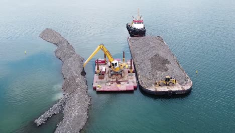 construcción de rompeolas con excavadora moviendo rocas de barcaza en el agua