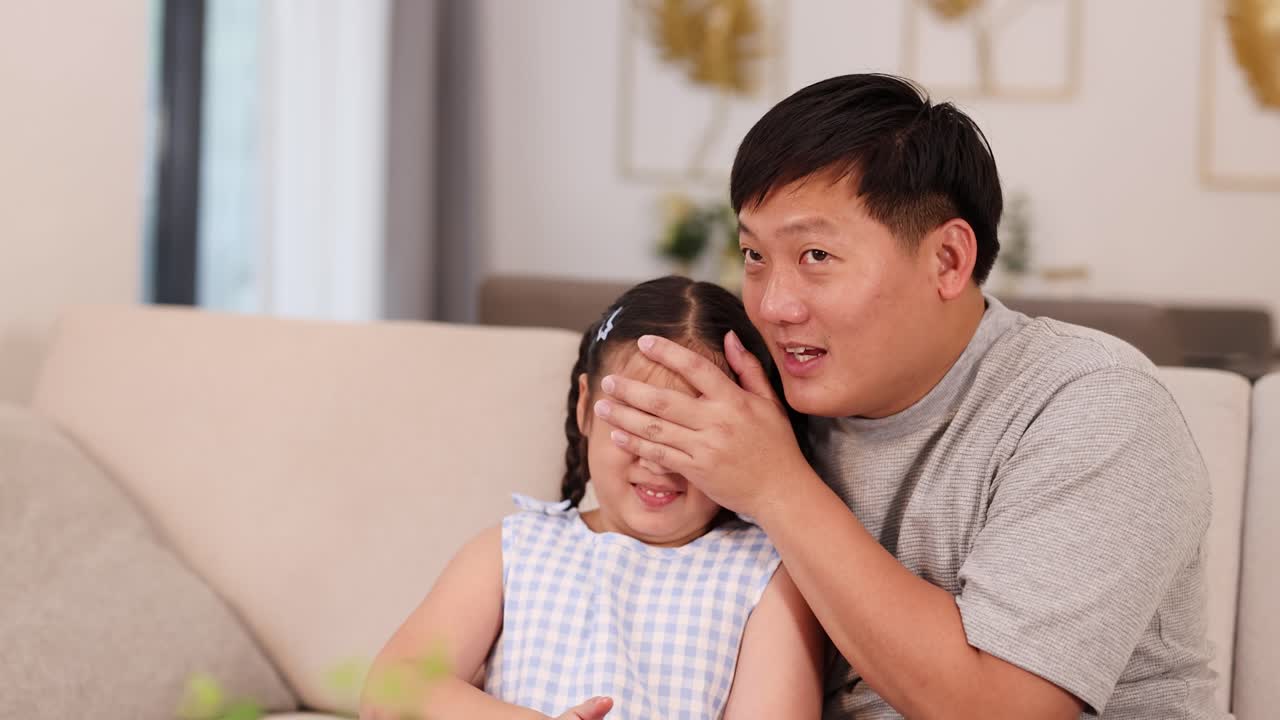 A father playfully covers his daughter's eyes while sitting on a cozy sofa in a warmly lit living room