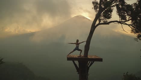 woman lunging into warrior pose on tree platform with moody mist and view of mount agung