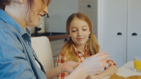 Young-mother-spreading-the-peanut-butter-on-the-bread-and-give-it-to-her-pretty-daughter-to-eat-in-the-modern-kitchen.-Close-up.-Indoor