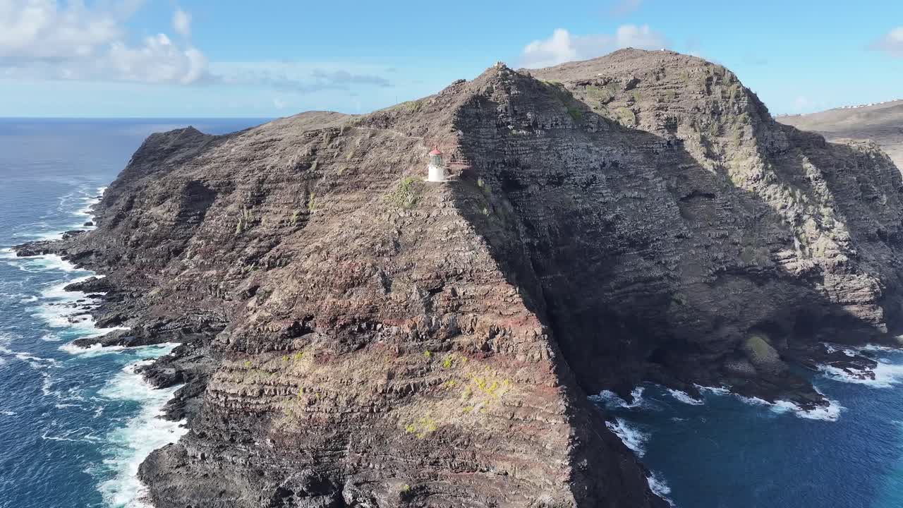 Aerial drone footage of a lighthouse perched on Oahu’s windward coast cliffs, Hawaii, showcasing dramatic rocky coastline, turquoise ocean waves, lush tropical vegetation, and panoramic ocean views