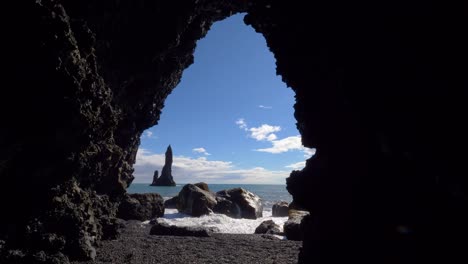 waves crash on large black rocks with stoic rock spire in ocean, black beach iceland