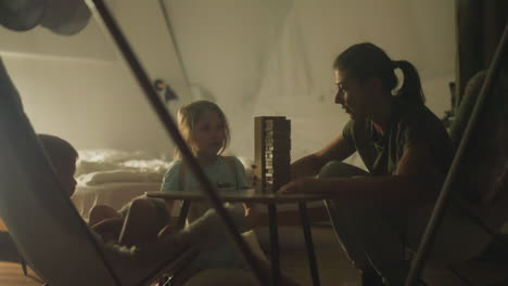 cheerful mother prepares wooden tower for playing jenga with son and daughter. family sitting at small table in cozy tourist chairs inside glamping camp in nature