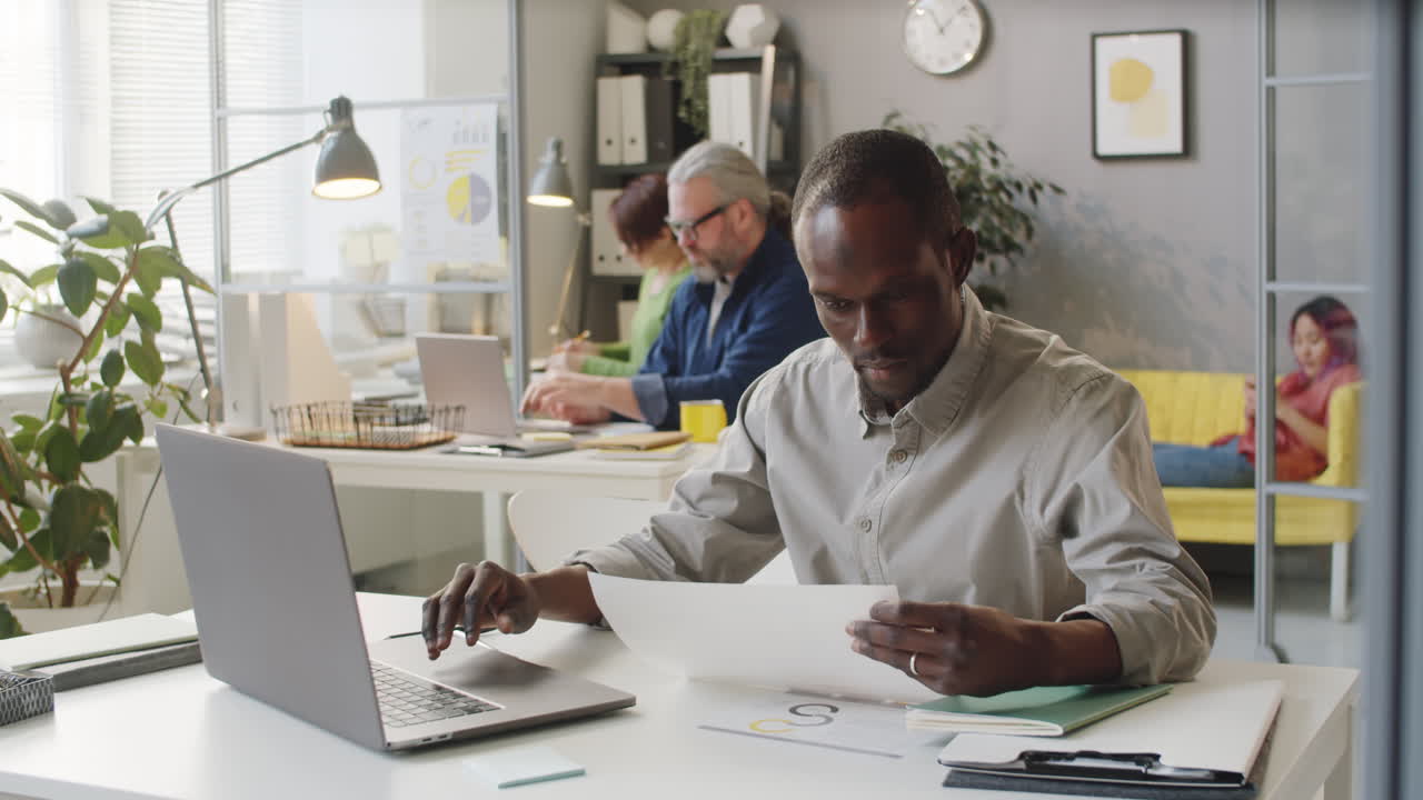Employé de bureau afro-américain utilisant un ordinateur portable et lisant un document