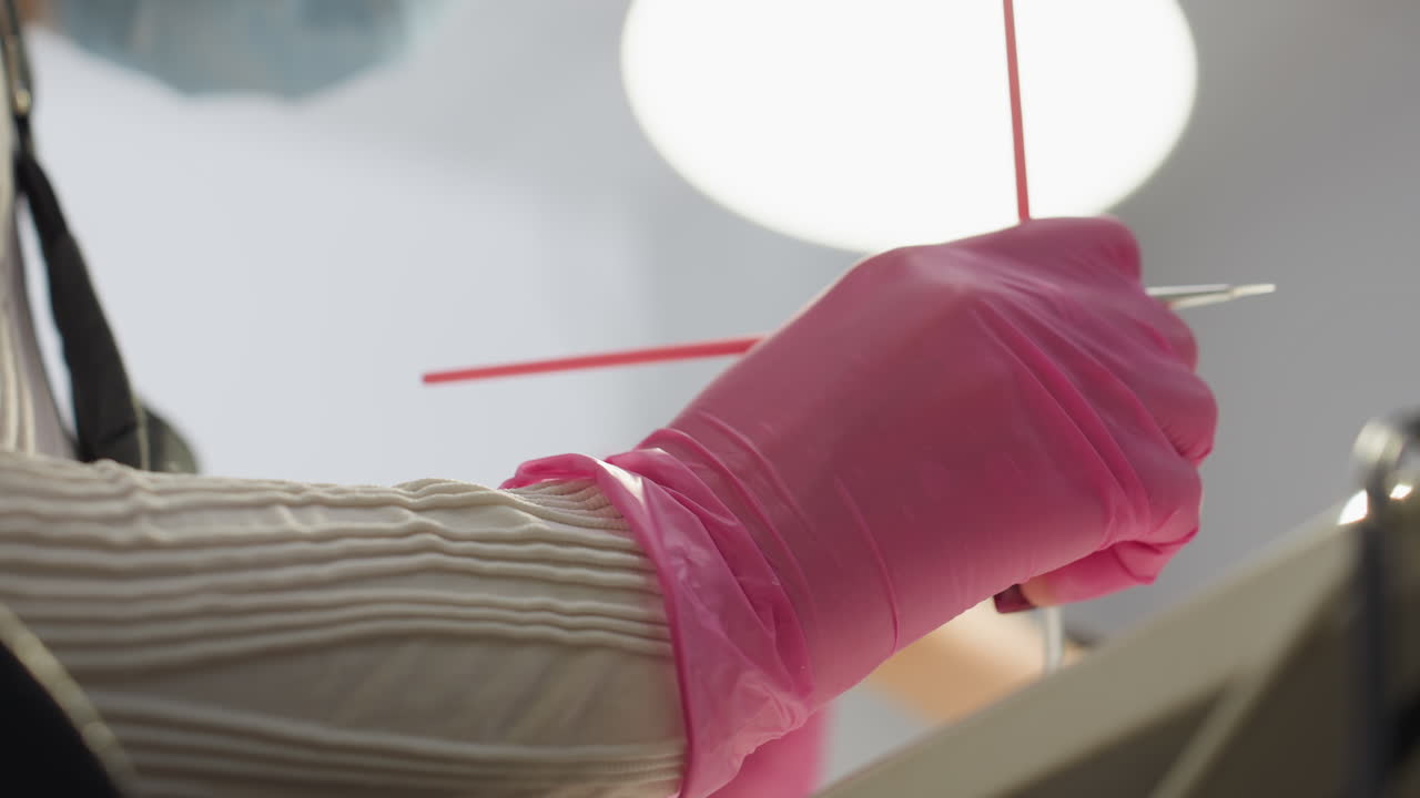 Nail technician wearing pink gloves dips fine brush into hardener and carefully applies product to client thumb under bright salon lamp, showcasing detailed nail enhancement process