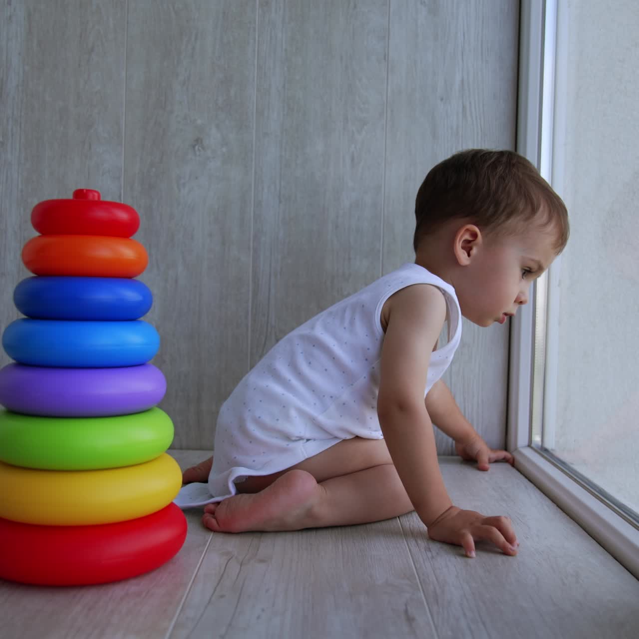 Lovely small kid in a white bodysuit sits at the window. Baby boy looks down attentively. Toy pyramid stands beside