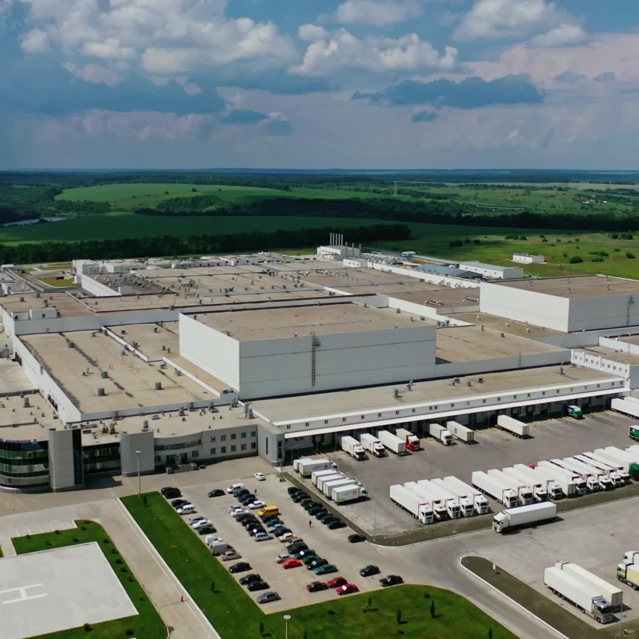 Manufacture zone in nature. Modern territory of huge industrial complex with car parking surrounded by green landscape. Aerial view. Camera moves back.
