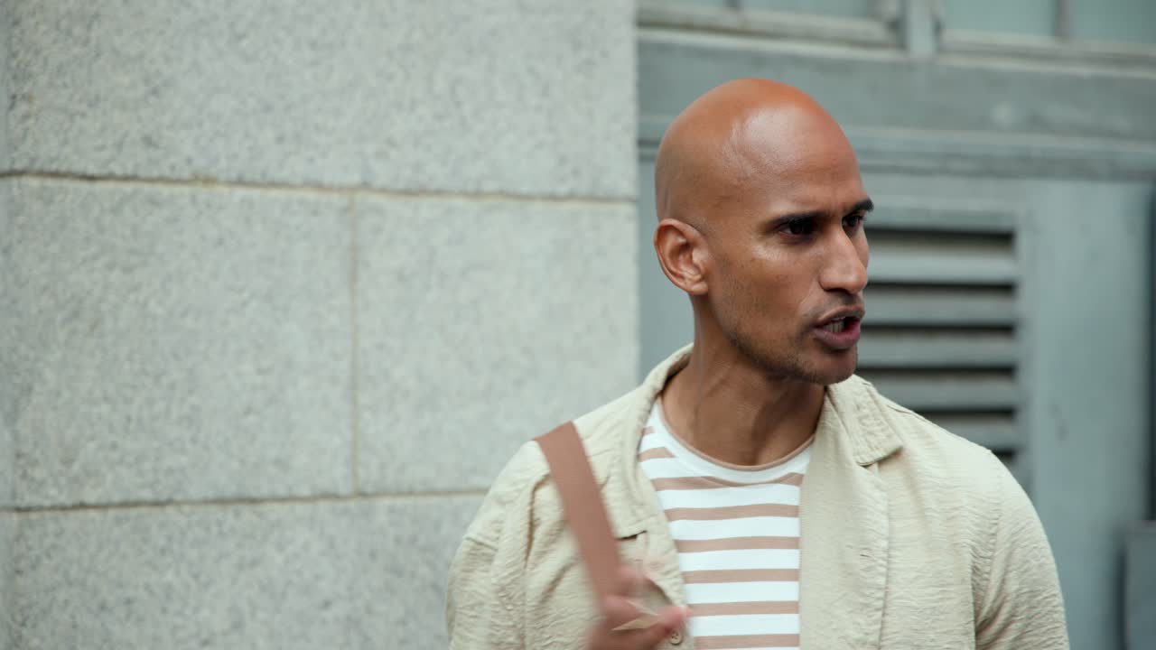 Man standing by wall, holding bag strap, in striped tee, speaking, pointing to smartphone appearing