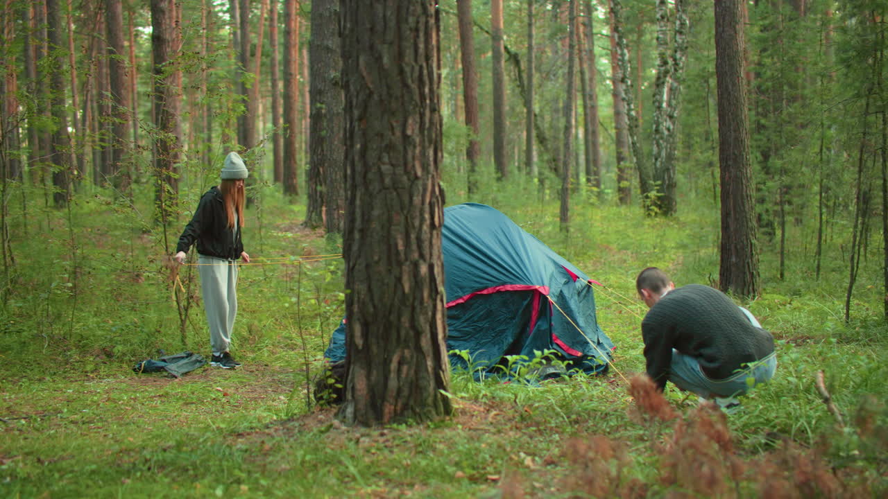 Back view of man crouched down hammering yellow rope into ground while woman holds and separates tangled lines during tent setup in forest, surrounded by trees and green vegetation