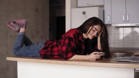 imágenes en cámara lenta de una joven sonriendo y riendo mientras se acuesta en la superficie de su mesa de la cocina y envía mensajes de texto a alguien.