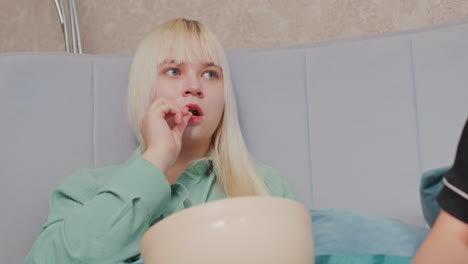 blonde girl in green shirt holds bowl and eats chips while resting on cushioned bed, partial view of another person beside her suggests shared moment during casual indoor relaxation or snack break