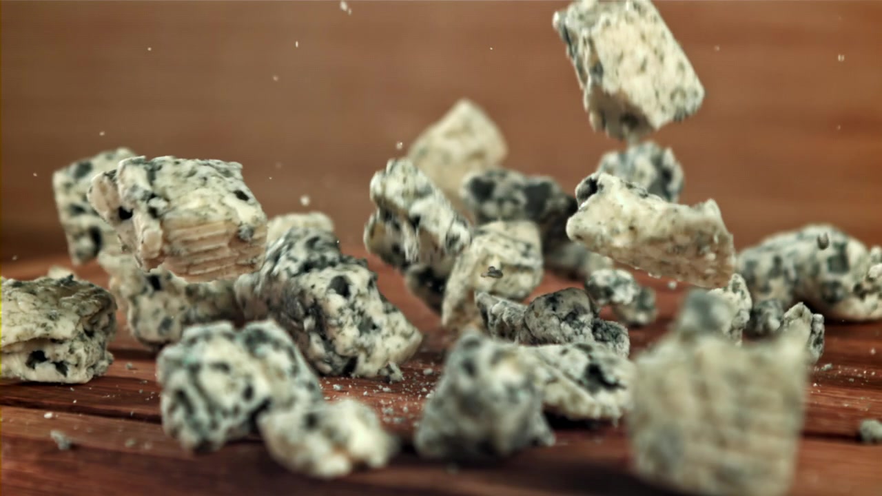 Pezzi di formaggio erborinato che cadono su un tagliere di legno al rallentatore.