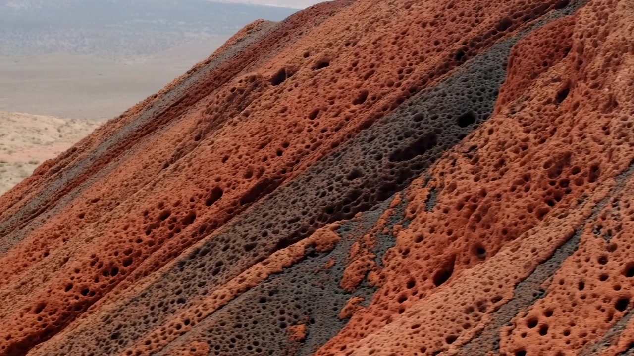 Vista obliqua ravvicinata-media che mostra un ripido pendio roccioso eroso con fitta alveolatura a nido d'ape, striature minerali diagonali color carbone, superficie ruvida a strati, toni terrosi e una forte texture naturale.