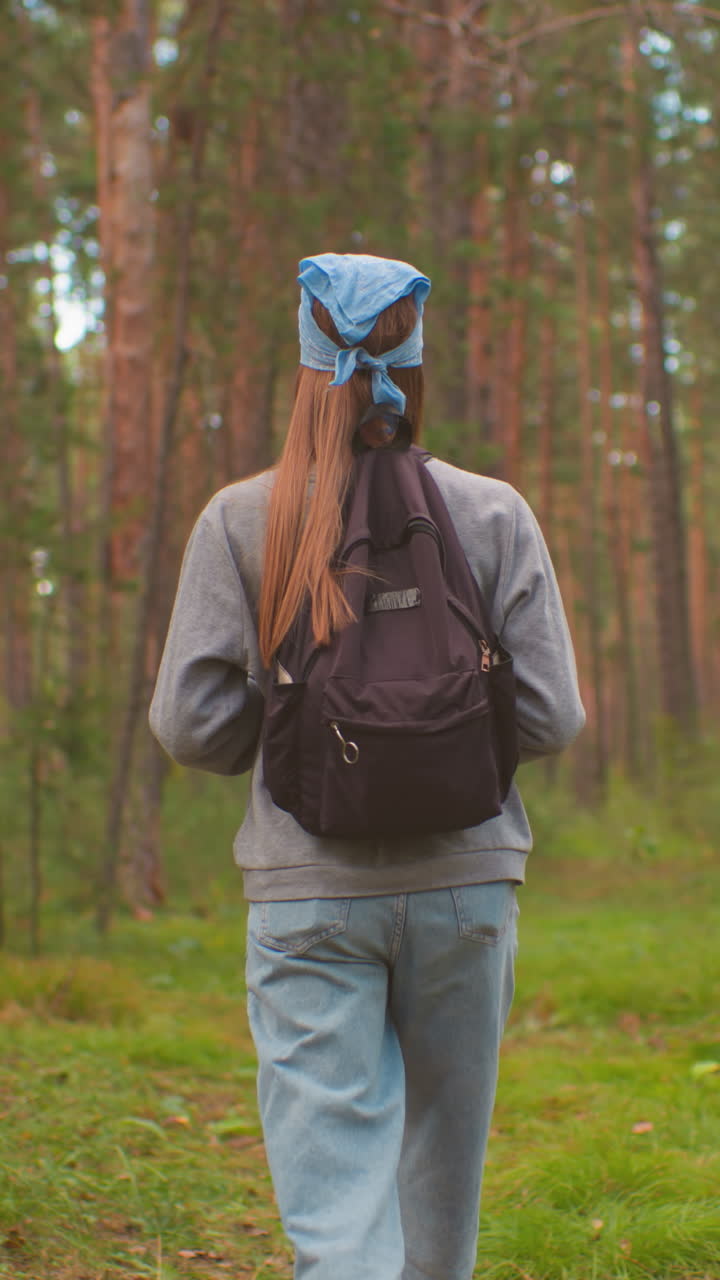 vista de la espalda de una mujer joven ajustando las correas de la mochila mientras camina a través de un camino pacífico y exuberante del bosque, con un bandana azul y rodeada de vegetación vibrante y árboles altos