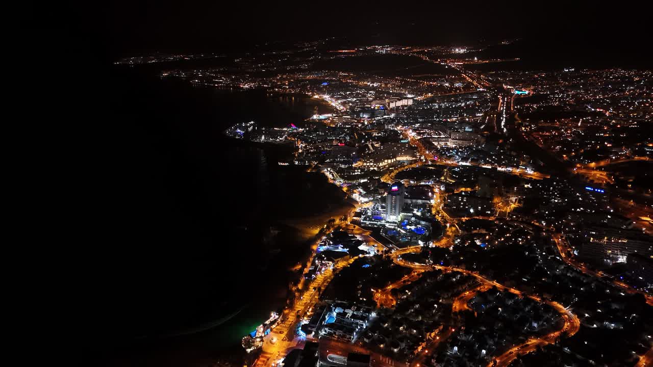 Nighttime drone view of a coastal city in Tenerife. Streetlights reflect on the Atlantic as the city glows quietly along the shore