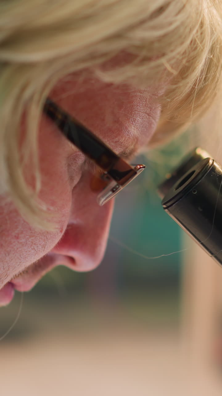 vista de cerca de un científico con una peluca rubia y gafas examinando intensamente una muestra a través de un microscopio en un laboratorio con un fondo borroso