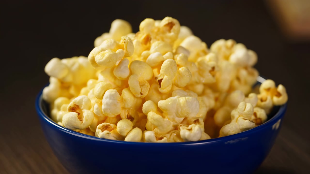 Scena ravvicinata in movimento di popcorn imburrati ammucchiati in una ciotola di ceramica blu scuro su un tavolo scuro, con popcorn, burro, snack, dettaglio ciotola, che mostra un primo piano, cibo, chicco, contesto giallo.