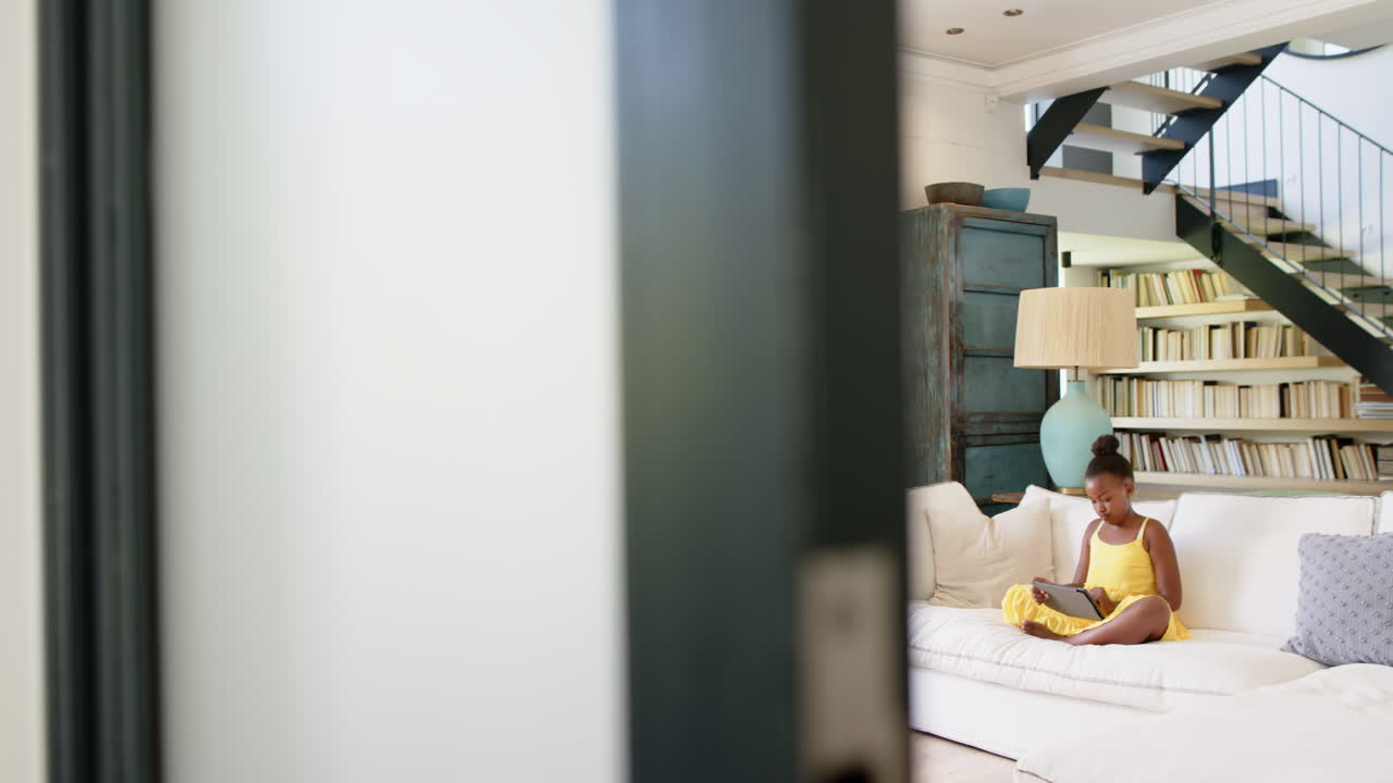 Sitting on couch, girl using tablet in cozy living room with bookshelves, copy space