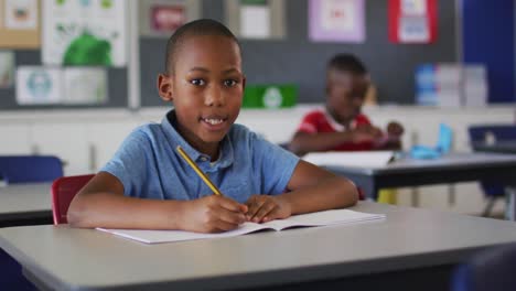 Retrato-De-Un-Feliz-Colegial-Afroamericano-Sentado-En-El-Aula,-Tomando-Notas,-Mirando-La-Cámara