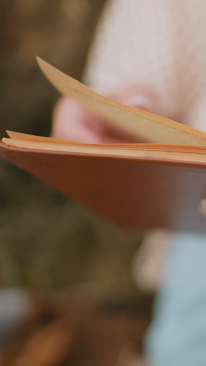 primer plano de manos sosteniendo un libro abierto, hojeando páginas, borrón suave en el fondo, con pantalones cortos azules y un momento de lectura relajado en un entorno tranquilo al aire libre