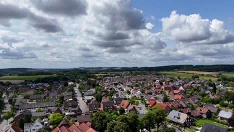 german village with traditional gabled houses, brick facades, tiled roofs and well-planned residential streets, surrounded by rolling farmland under dramatic sky. aerial rising wide shot