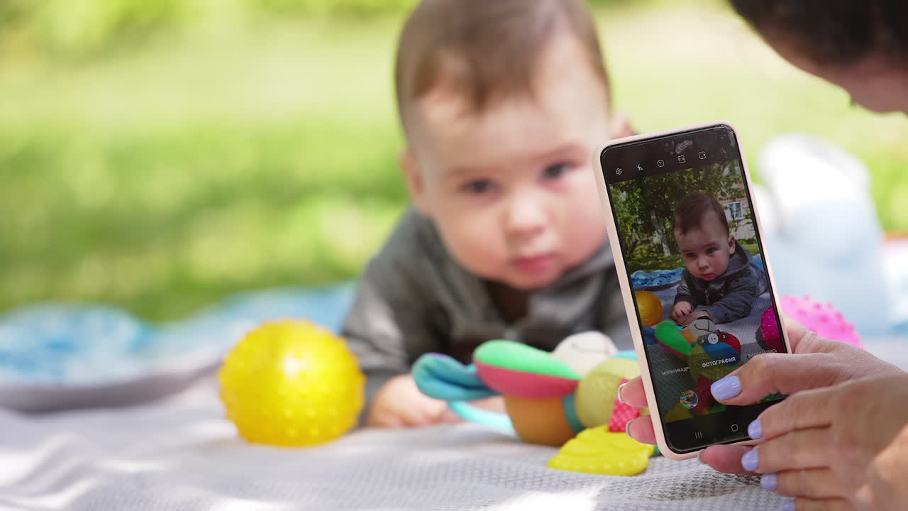 Mom's hand is taking pictures of her baby on the phone. Adorable boy lies peacefully posing. Blurred backdrop.