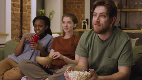 three roommates are watching a movie while eating popcorns and potato chips sitting on sofa at home