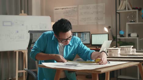 asian man engineer with a laptop measuring blueprint at the office