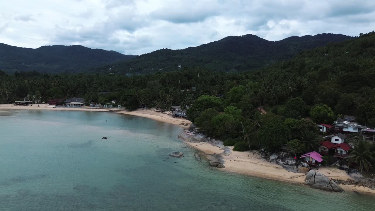 Aerial drone footage of Koh Phangan on a cloudy day, showing tropical coastline, beachfront bungalows and scenic mountains and hills in the background of southern Thailand