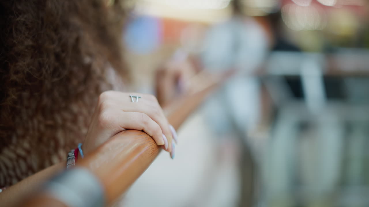 Close-up of a hand resting on an iron railing, adorned with a chain bracelet and red rope, the hand's delicate, polished nails, while the blurred background features people moving in a busy setting
