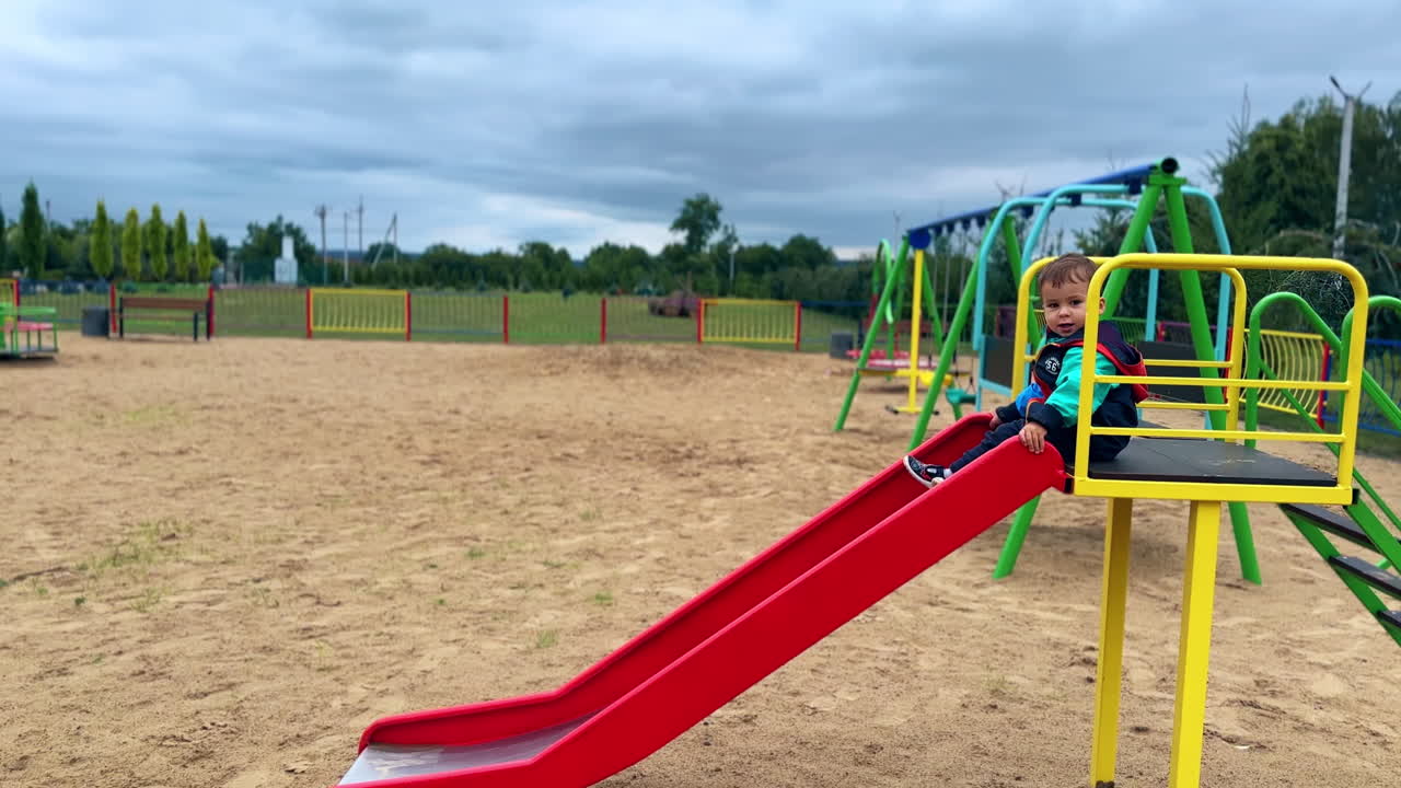 Lovely baby boy sits on the slide. Kid goes down and sits looking around.