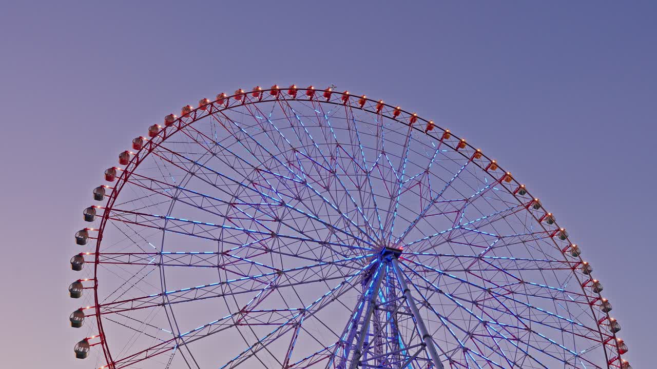 The upper half of the Diamond and Flower Ferris Wheel illuminated with blue lights against a pastel twilight sky