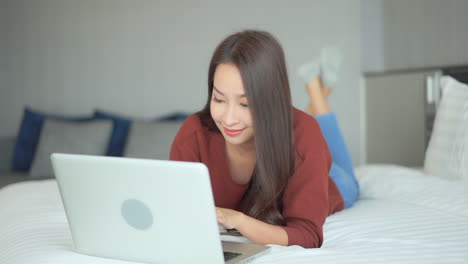 a pretty young woman in jeans and a sweater lies on a big comfortable hotel bed as she successfully completes an online interaction