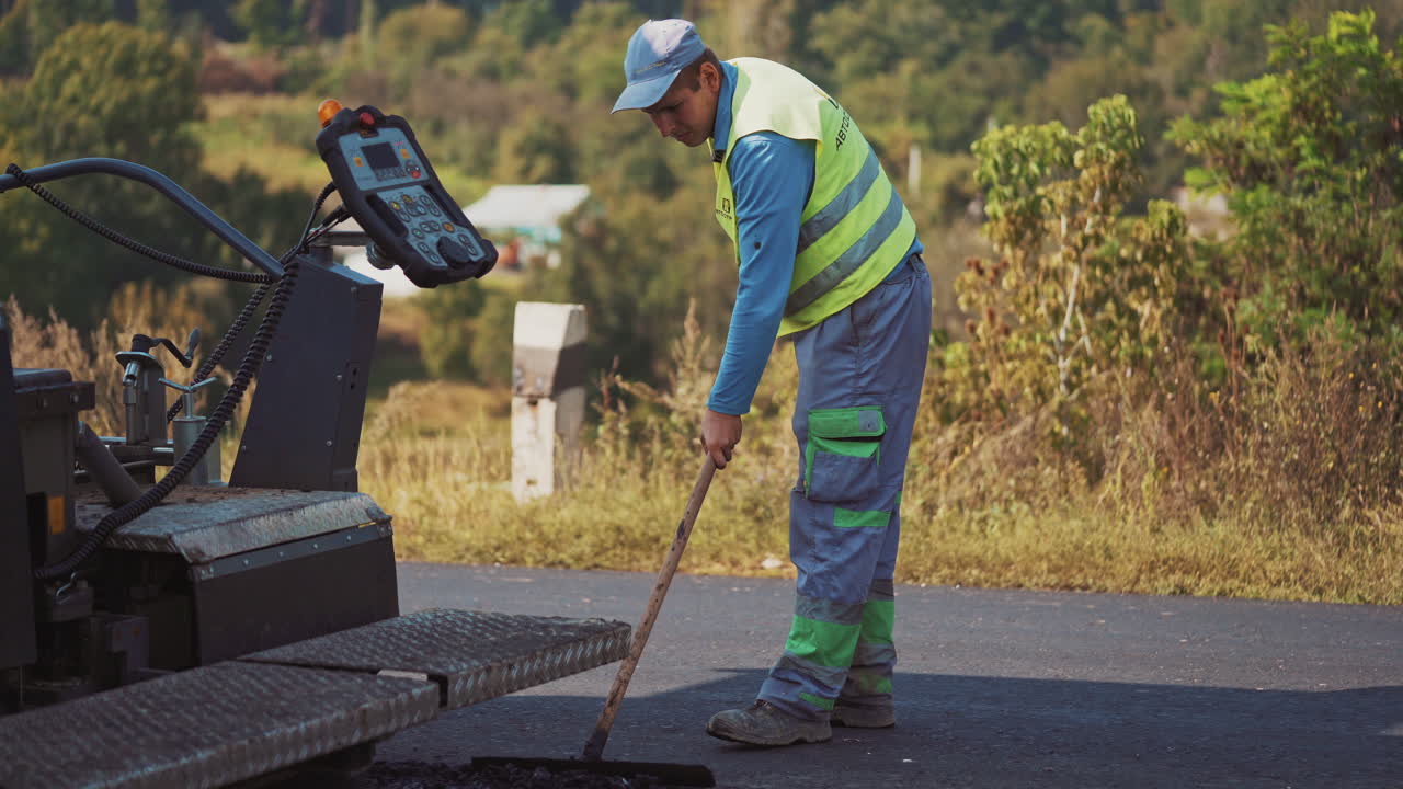 Close-up part of asphalt paver and a worker. Worker man in protective clothing works with special instrument after paver machine on the road.